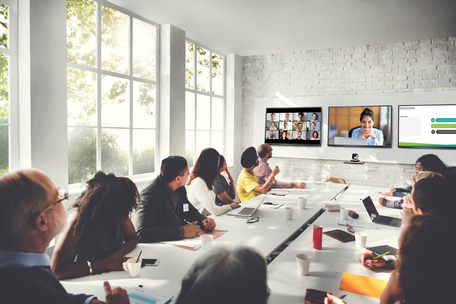 A modern meeting room with a large conference table, three displays in the front, and people participating in a video conference.