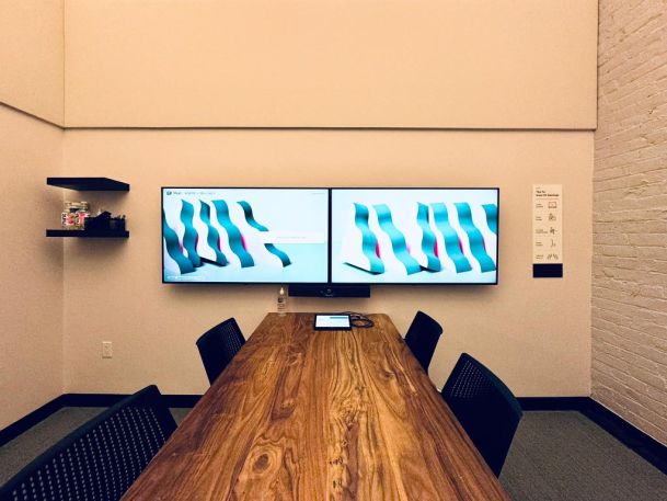 Conference room with a wooden table, four chairs, and dual wall-mounted displays showing abstract visuals.