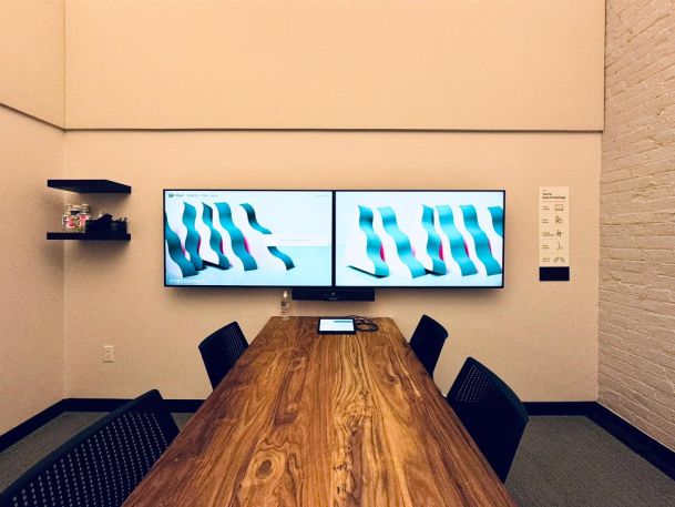 Conference room with a wooden table, four chairs, and dual wall-mounted displays showing abstract visuals.