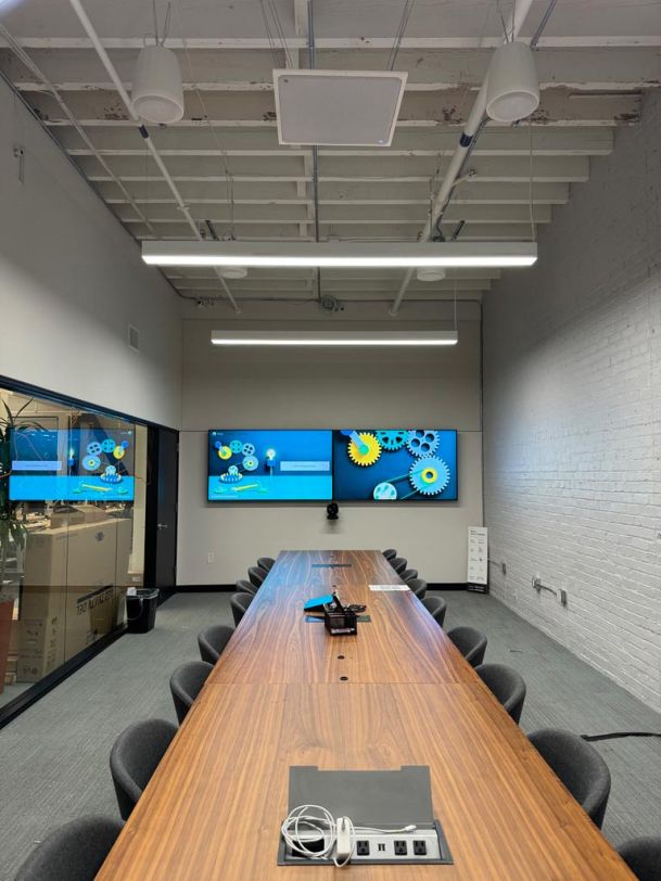 Long conference table with eight chairs in a loft-style room, featuring dual wall-mounted displays and exposed ceiling beams.