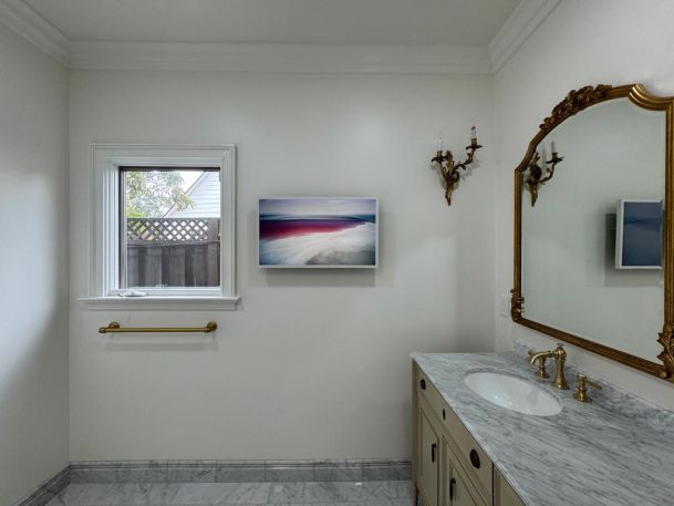 A white bathroom with a marble countertop, featuring a large mirror, a decorative light fixture, and a landscape painting.