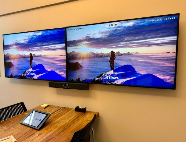 A close-up of two large wall-mounted screens showing a person on a mountain peak above a cloudscape.