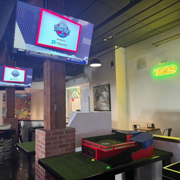 Bar with sports on multiple wall-mounted TVs, featuring a mini-golf putting area in the foreground.