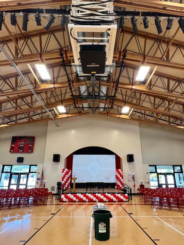 A school gymnasium with a stage decorated with red and white balloons, facing a screen ready for a presentation.