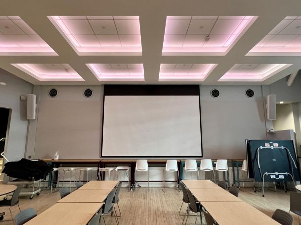 Spacious presentation room with rows of tables and a large projection screen under soft pink lighting.