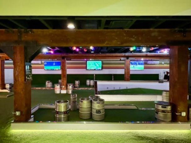 An indoor recreational space with keg barrels, a mini-golf area, and multiple screens displaying sports.