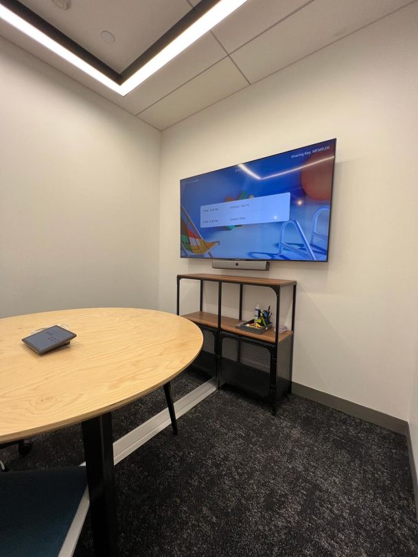 A small meeting room with a round table, a wall-mounted screen, and a storage shelf below.