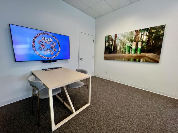 Minimalist meeting room with a small wooden table, two chairs, and a wall-mounted display, alongside wall art.