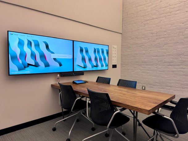 A modern meeting room with a wooden table, black chairs, and dual wall-mounted screens displaying a blue pattern.