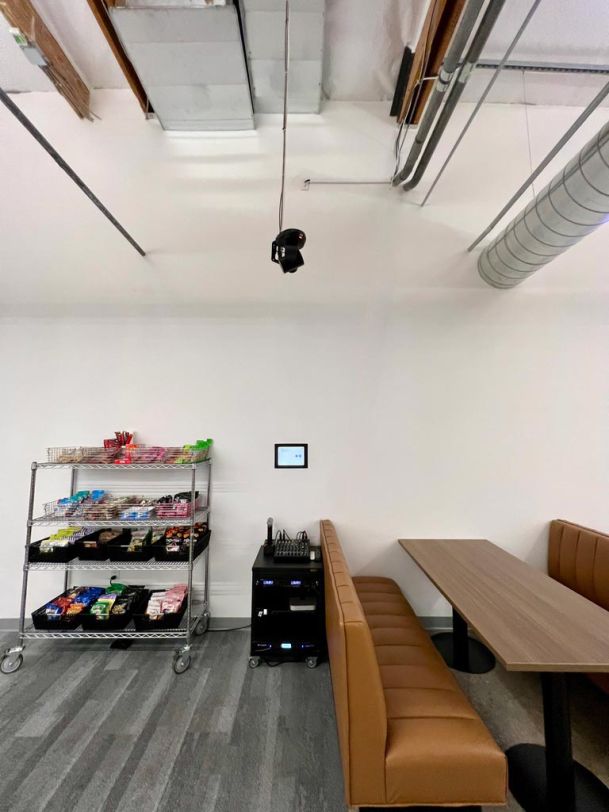 A snack area with a bench, a table, and a cart with various snacks, under an exposed ceiling with visible pipes.