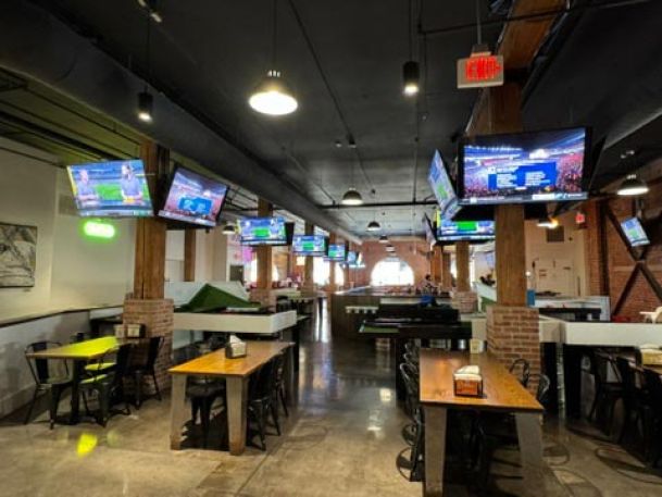 Spacious bar with wooden pillars, multiple wall-mounted TVs showing sports, and several tables arranged for dining.