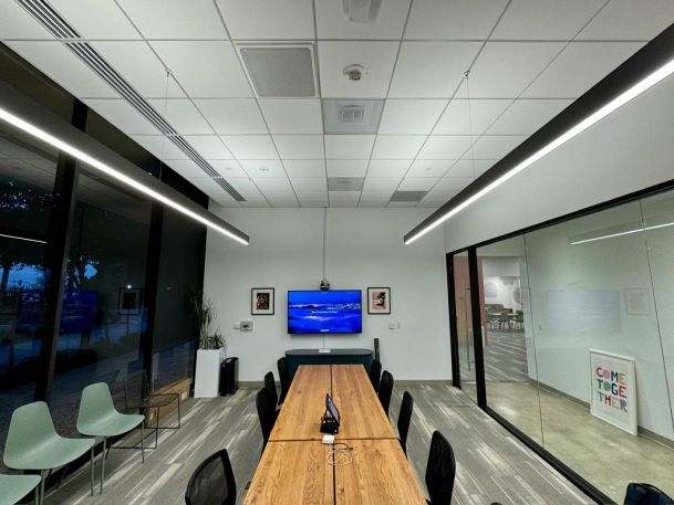 A sleek meeting room with a long wooden table, a TV screen at the end, and large windows showing an outside view.