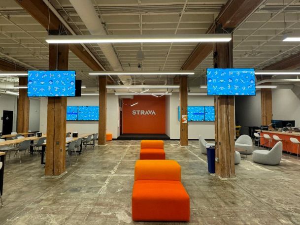 Open office space with orange lounge seating, multiple wall-mounted screens, and an exposed industrial ceiling.