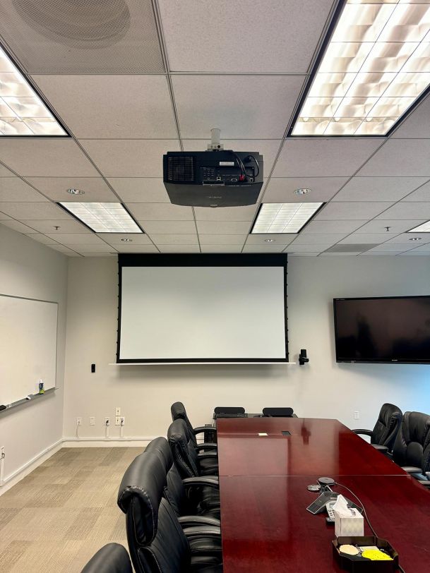 A conference room with a large table and leather chairs, featuring a projector and a white screen for presentations.