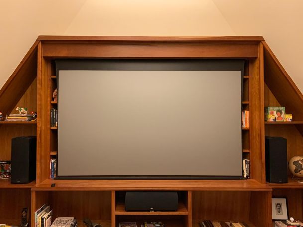 A large projection screen is set in a wooden frame flanked by shelves, within a home theater setup with visible speakers and books.