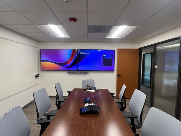 Conference room with six chairs around a wooden table, featuring dual wall-mounted displays and a video conferencing setup.