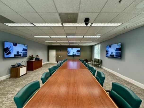 Large conference room with a long wooden table, green chairs, and three wall-mounted displays for presentations.