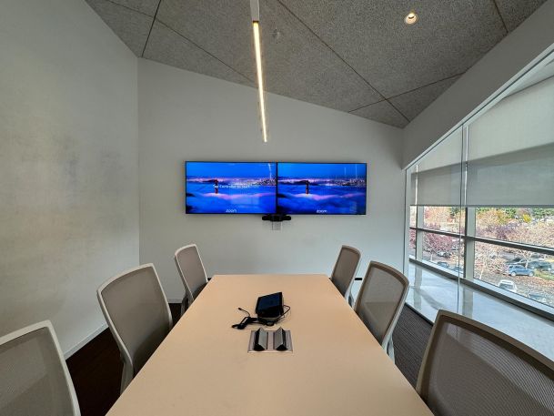 A small meeting room with a table and chairs, featuring dual wall-mounted screens displaying a scenic image.