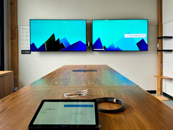 Meeting room with a large wooden table, two wall-mounted screens, and a tablet on the table for collaboration.