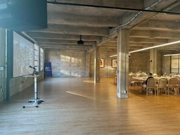 A large industrial-style room with concrete walls, wooden floors, and a dining table set for an event, with a projector and microphone stand in view.