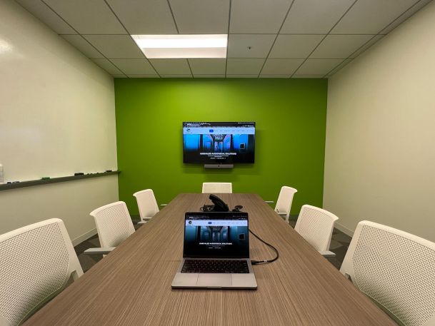 A small conference room with a table, chairs, a laptop on the table, and a wall-mounted TV displaying a website, framed by a green accent wall.