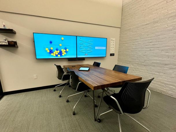 A meeting room with a wooden table, four chairs, and dual wall-mounted screens showing a colorful graphic with shapes.