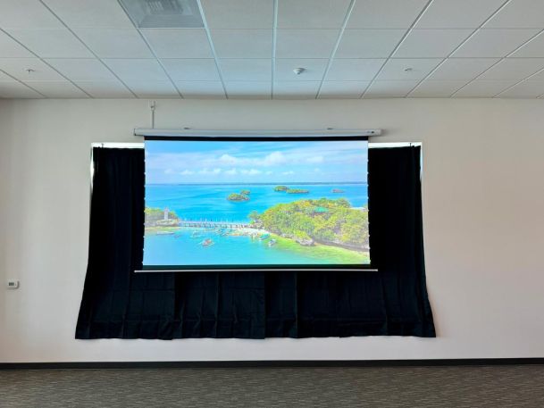 A projector screen displaying a vibrant beach view, framed by black curtains in a room with a tiled ceiling.