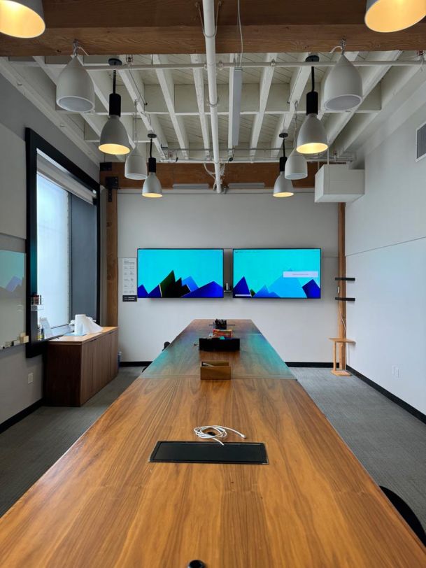 A meeting room with wooden ceiling beams, pendant lights, a long table, and dual wall-mounted screens showing a blue graphic.