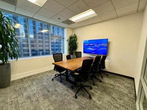 Bright conference room with a large wooden table, black chairs, and a wall-mounted display overlooking large windows.