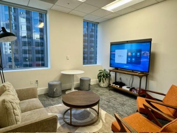 A cozy lounge area with a TV displaying a Zoom screen, surrounded by chairs, tables, and city views from the windows.