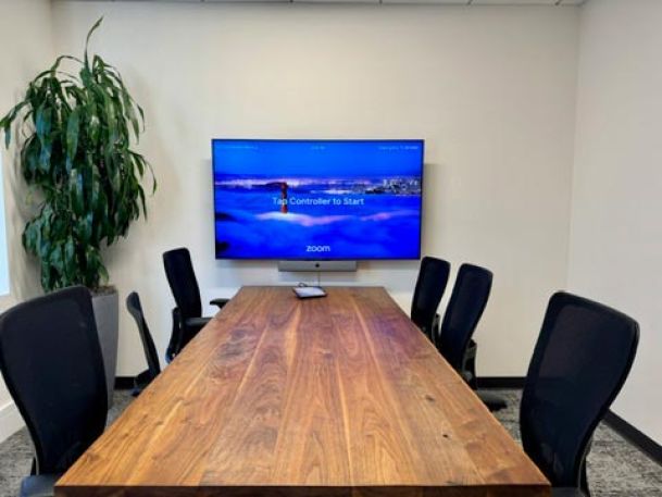 A small meeting room with a wooden table, surrounded by chairs and a wall-mounted screen displaying a Zoom setup.