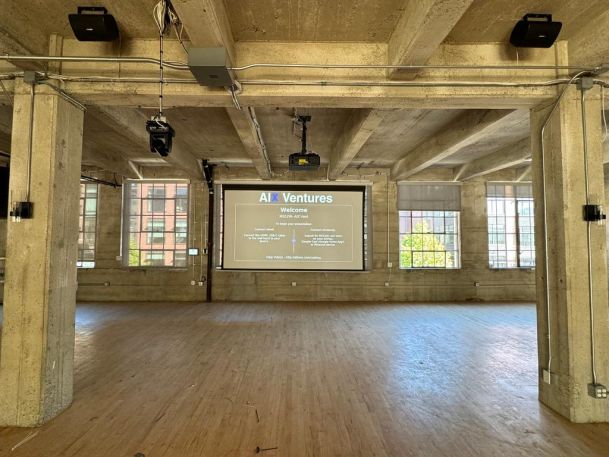 A spacious industrial room with a wooden floor and large windows, featuring a projection screen displaying a welcome message, under a high concrete ceiling.