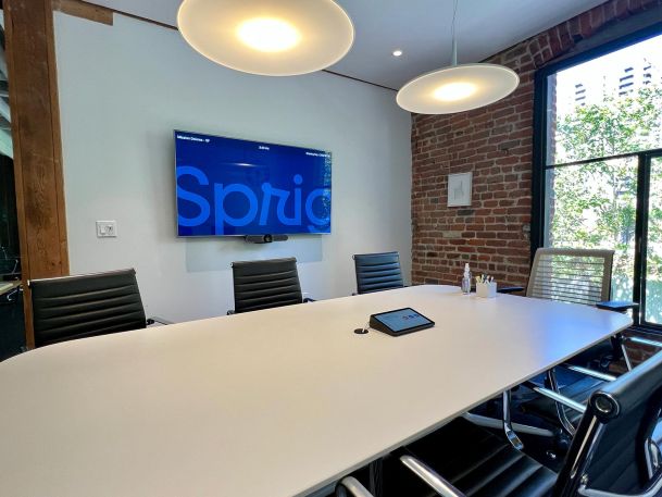 Small meeting room with a white conference table, black chairs, and a wall-mounted display screen.