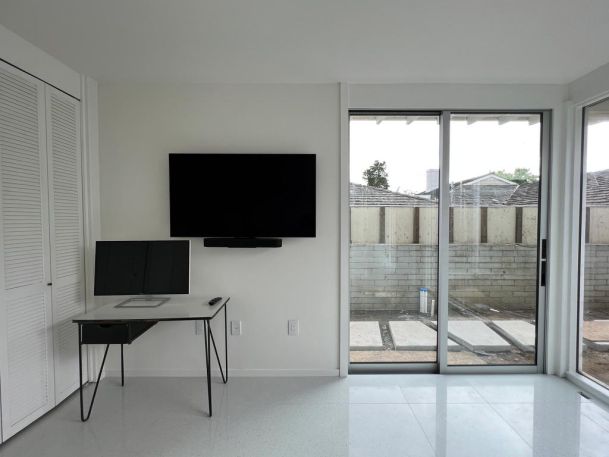 A minimalistic office with a desk, a monitor, and a wall-mounted TV near sliding glass doors.